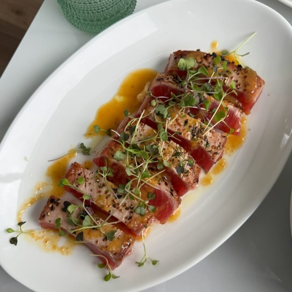 Seared tuna slices arranged on a white plate with microgreens, sesame seeds, and orange sauce.