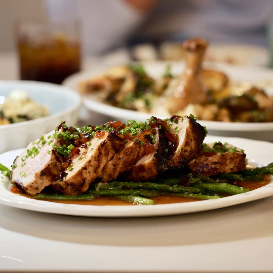Sliced grilled chicken breast topped with herbs and sauce, served over asparagus on a white plate.