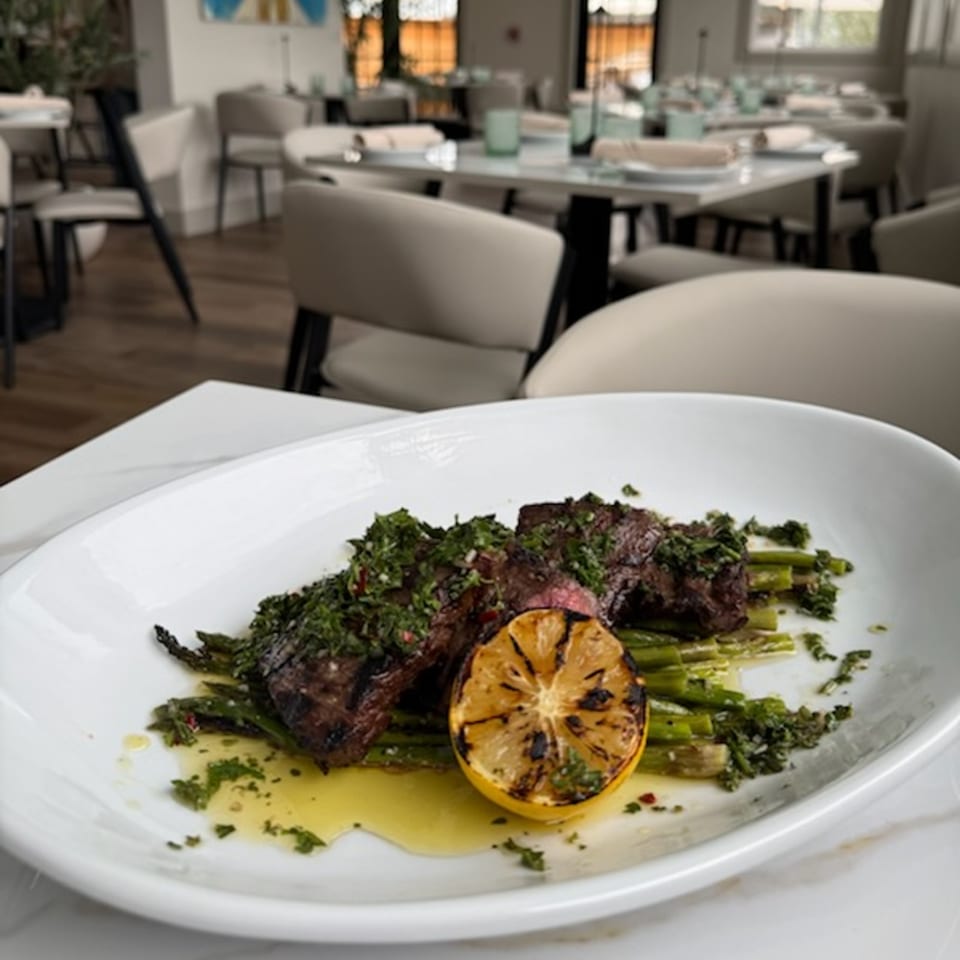 Grilled steak with green vegetables, herbs, citrus, and sauce served on a white plate in an elegant restaurant dining room.