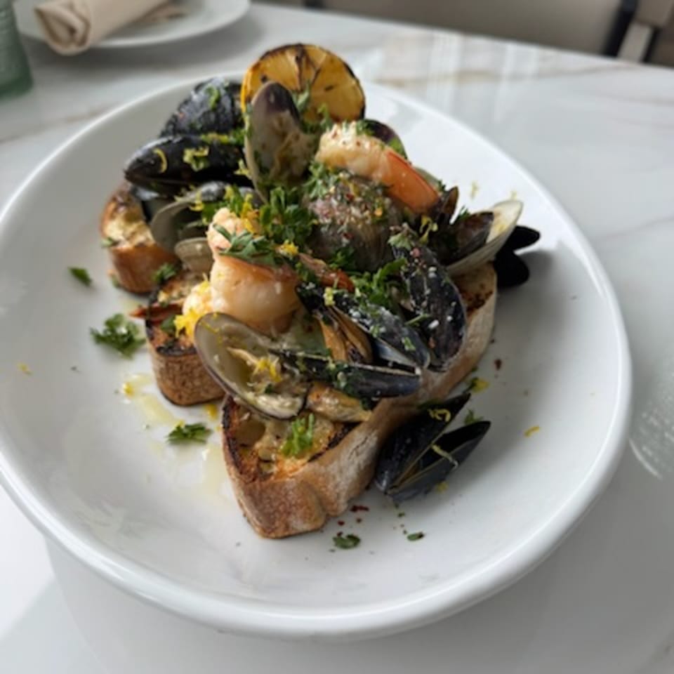 Platter of grilled mussels, shrimp, vegetables, herbs, and toasted bread served on a white plate.