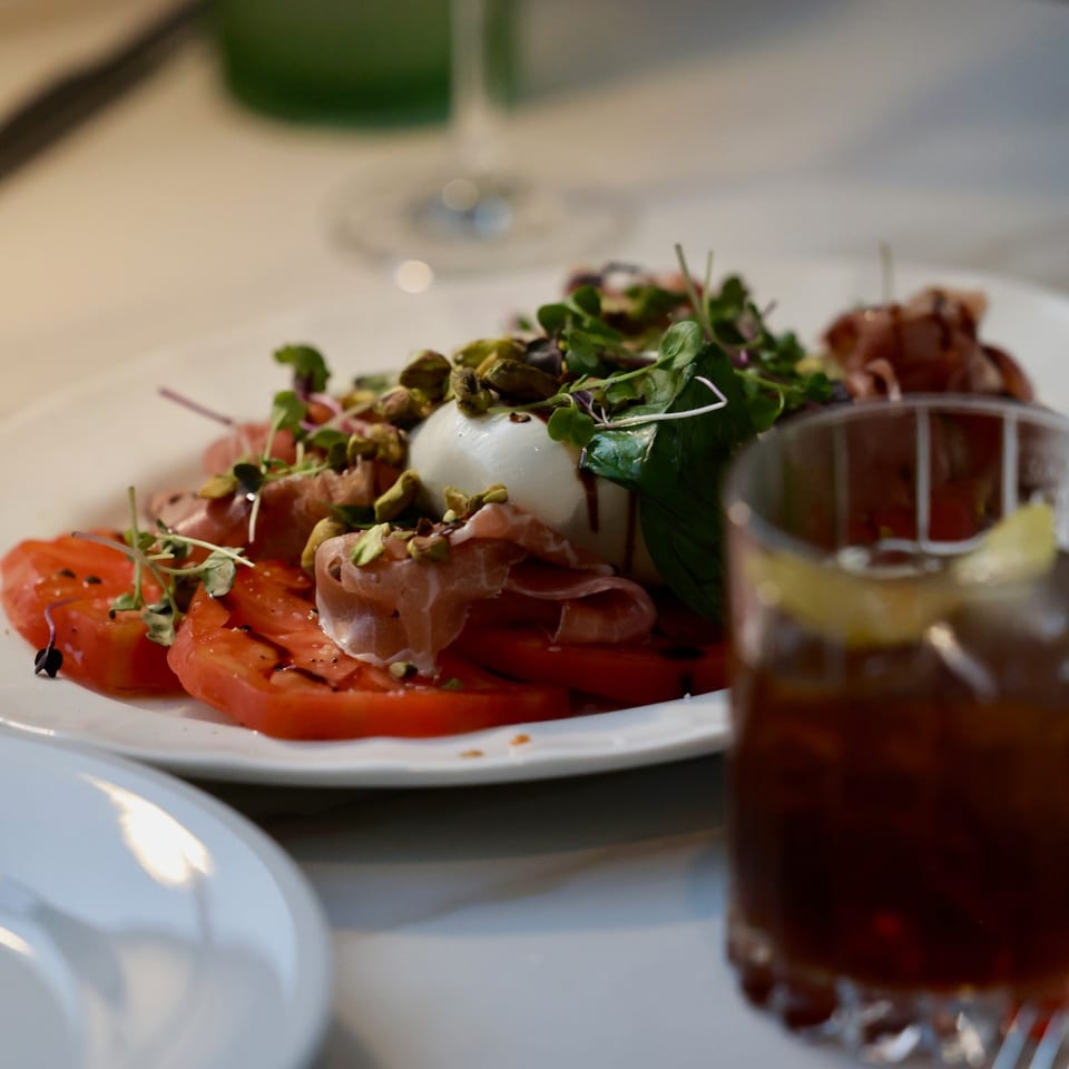 Gourmet plate of sliced tomatoes, cured meat, burrata, pistachios, and microgreens beside a blurred glass of red wine.